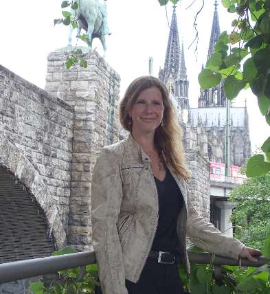 Porträt von Astrid Schwikardi Porträt einer Frau mit langen, welligen Haaren, lächelnd vor einer historischen Steinmauer.