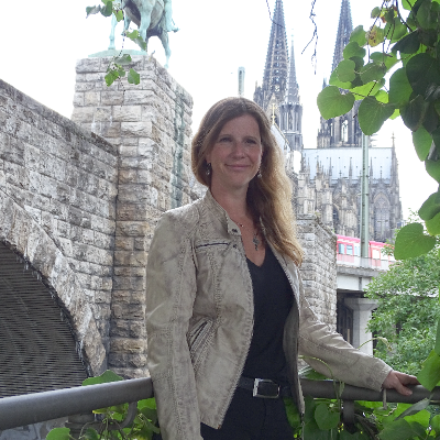 Porträt von Astrid Schwikardi Porträt einer Frau mit langen, welligen Haaren, lächelnd vor einer historischen Steinmauer.
